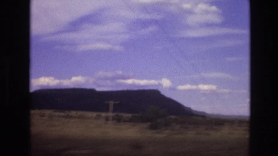1973 New Mexico travel footage frame with a flat-topped mesa, dry scrubland, distant roadside structures, and scattered clouds.