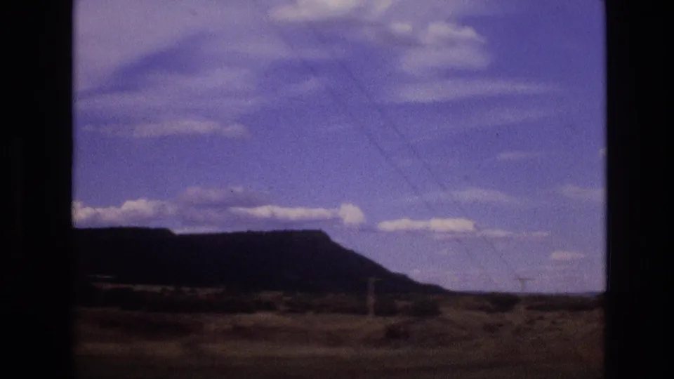 1973 New Mexico home-movie view of a dark mesa ridge across open desert terrain with low distant structures and a clouded sky.