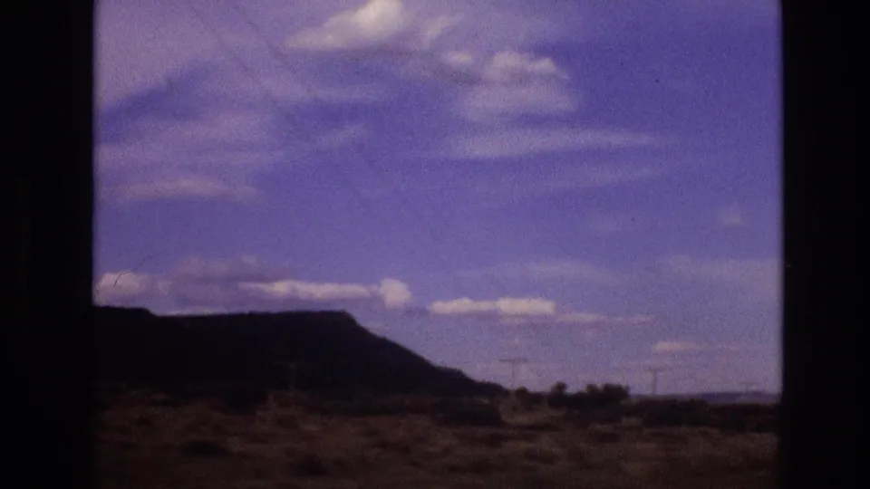 1973 New Mexico frame showing a dark mesa silhouette, sparse arid foreground, distant utility lines, and a bright cloud-streaked sky.