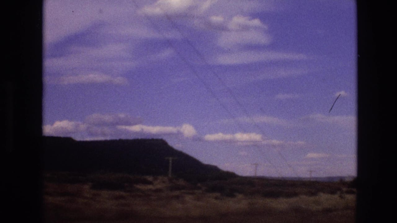 Dark-framed desert view with a rocky mesa, utility poles, blue sky, and scattered clouds.