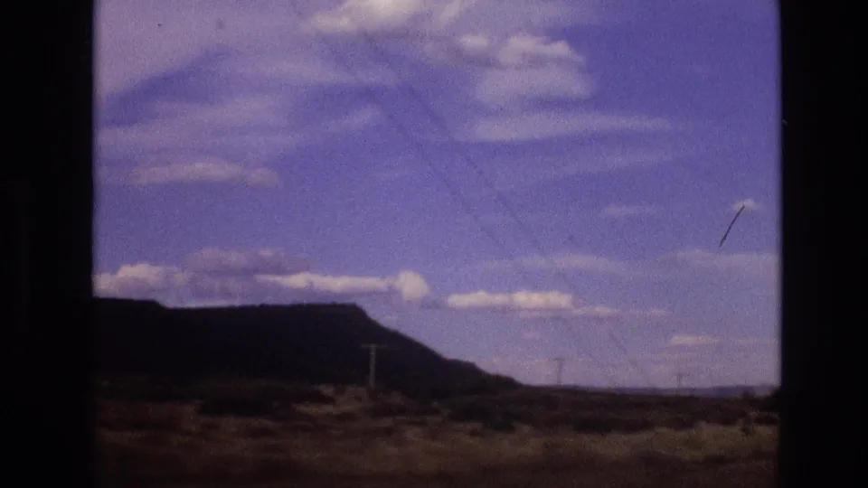 1973 New Mexico 8mm frame showing a dark window-framed desert landscape with a low rocky mesa, utility poles, scrubland, blue sky, and scattered clouds.