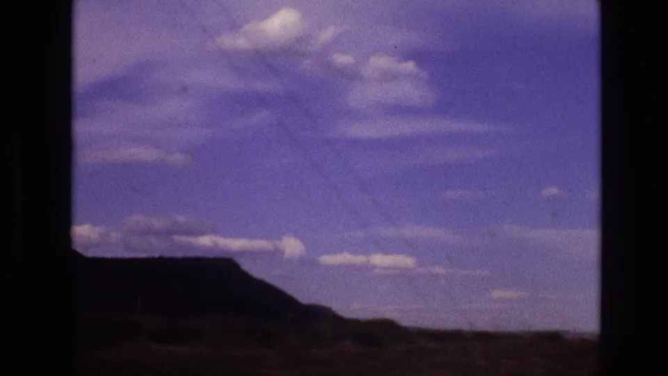 1973 New Mexico 8mm frame showing a low dark mesa across arid scrubland, with open blue sky, scattered clouds, and a dark window edge at the side.