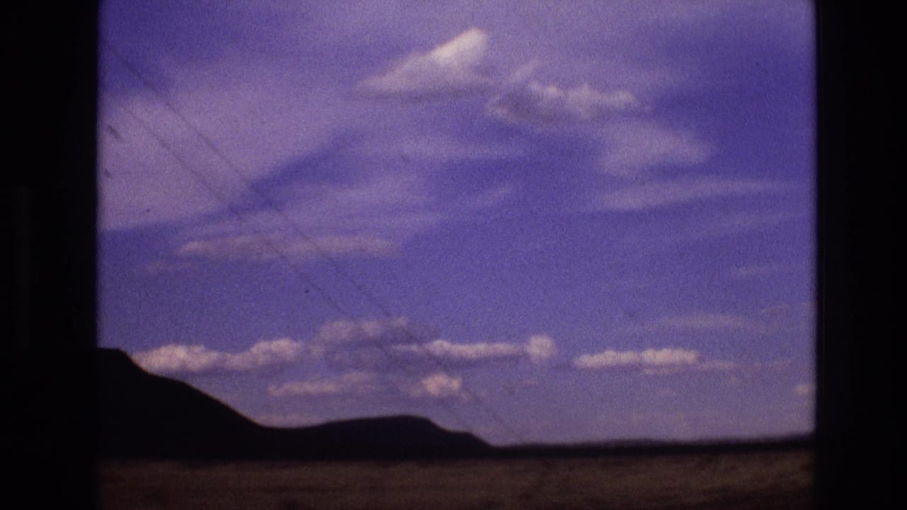 1973 New Mexico road-view frame with a low ridge, open desert foreground, purple-tinged sky, scattered clouds, and visible film gate edges.