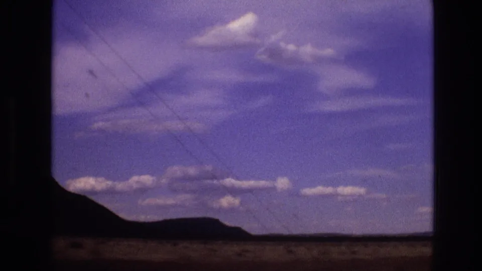 1973 New Mexico 8mm frame showing low desert hills, a flat foreground, scattered clouds, and the soft color-shifted look of a moving home-movie shot.