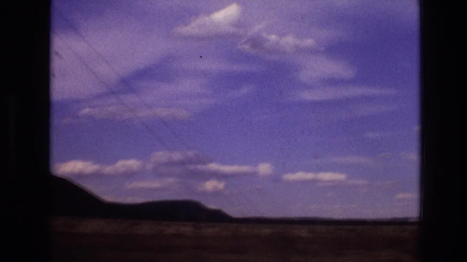 1973 New Mexico home-movie frame from a moving vehicle, showing a dark ridge line, open desert ground, scattered clouds, and faint scan scratches or film marks.