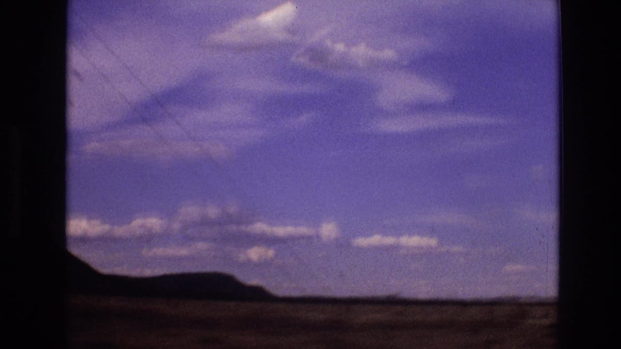 1973 New Mexico home-movie frame with low distant hills, open rural ground, and a purple-blue sky with scattered clouds.