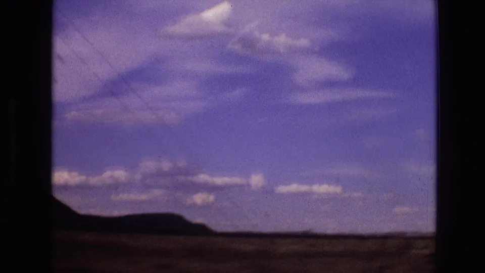 1973 New Mexico home-movie frame with low distant hills, open rural ground, and a purple-blue sky with scattered clouds.