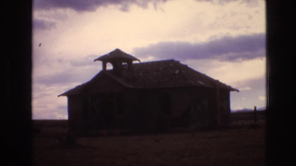 1973 New Mexico 8mm frame showing a weathered abandoned schoolhouse-like building with a small bell tower, dark openings, and open rural ground.