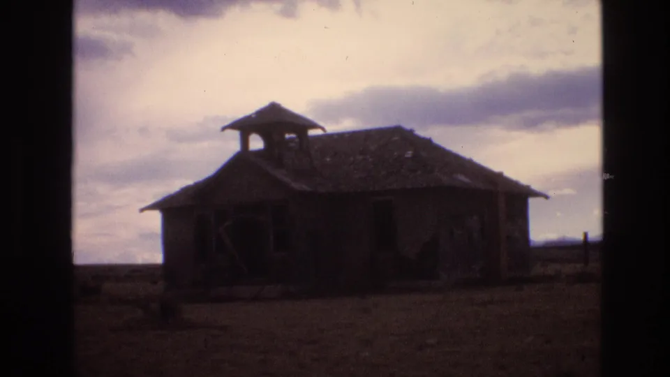 1973 New Mexico home-movie frame of a weathered single-story schoolhouse-like structure with a bell tower, set against open ground and a cloudy sky.
