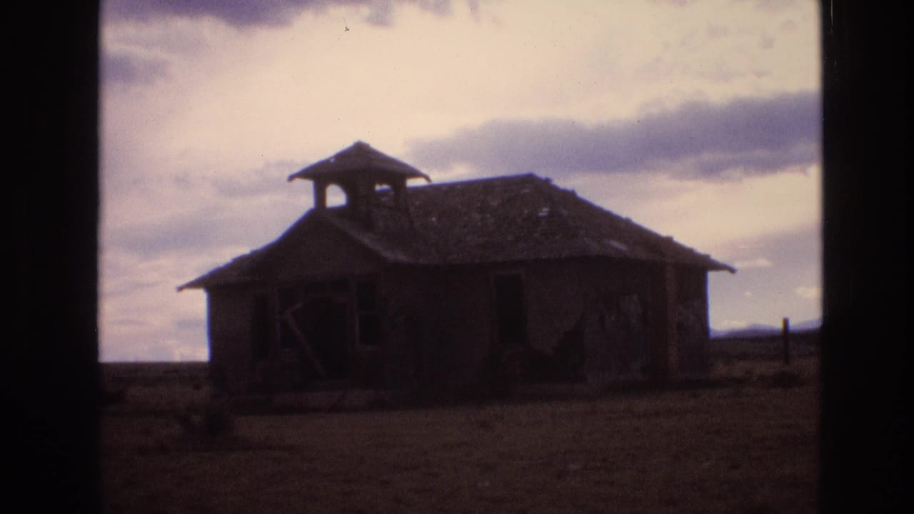 1973 New Mexico 8mm frame showing a dark, weathered schoolhouse or shed with a damaged roof standing alone on a rural plain under clouds.