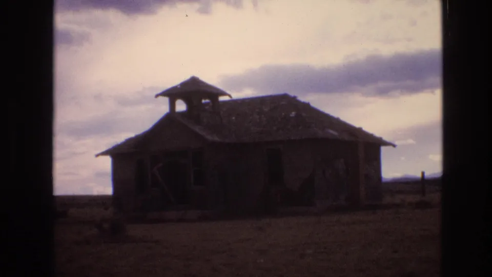1973 New Mexico 8mm frame showing a dark, weathered schoolhouse or shed with a damaged roof standing alone on a rural plain under clouds.