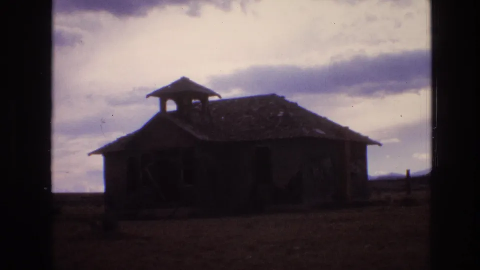 1973 New Mexico 8mm frame showing a closer handheld view of the abandoned schoolhouse or shed, with a dark facade and cloudy sky behind it.