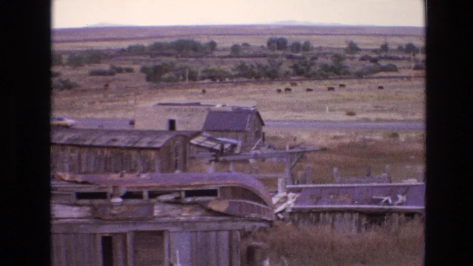 1973 New Mexico 8mm frame widening to abandoned farm buildings, fence lines, open pasture, distant cattle, and a low tree line on the rural plain.