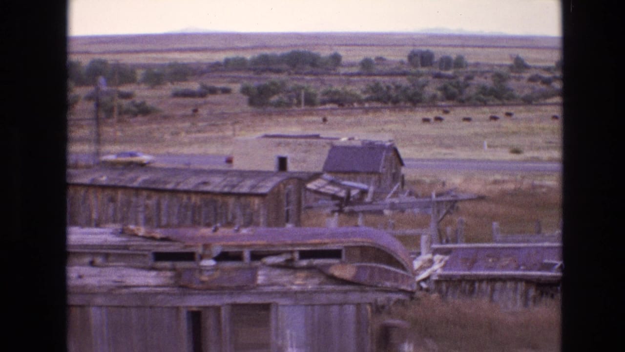 1973 New Mexico 8mm frame showing abandoned wooden railroad buildings with rusted metal roofs, dry grass, a parked car, distant cattle, and desert plains.