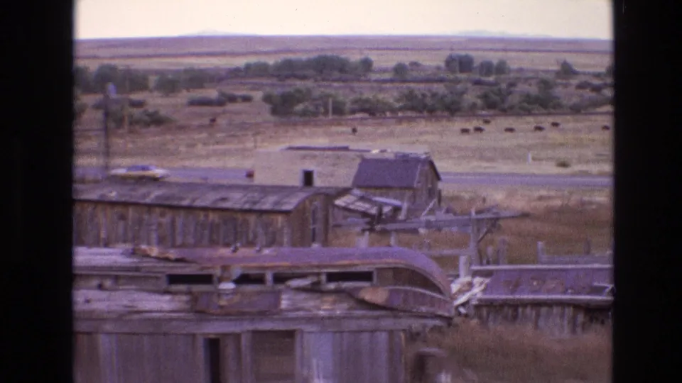 1973 New Mexico 8mm frame showing abandoned wooden railroad buildings with rusted metal roofs, dry grass, a parked car, distant cattle, and desert plains.