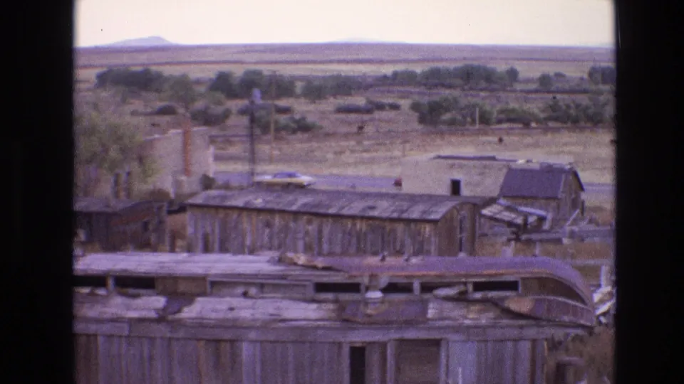 1973 New Mexico home-movie frame with deteriorating railroad structures, a pale parked car, utility poles, grazing cattle, and a low desert horizon.