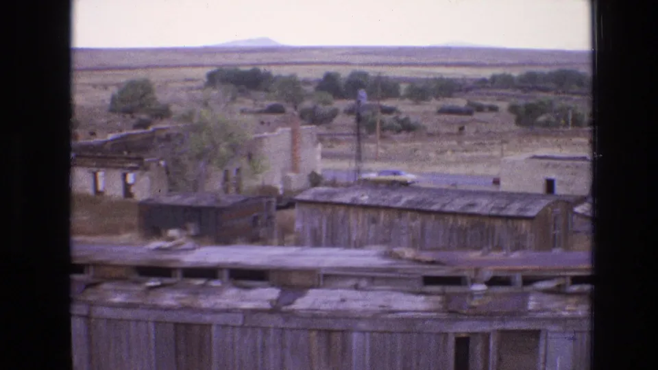 1973 New Mexico 8mm frame showing worn wooden buildings, roof edges, scattered trees, dry grass, distant cattle, and expansive desert land beyond.