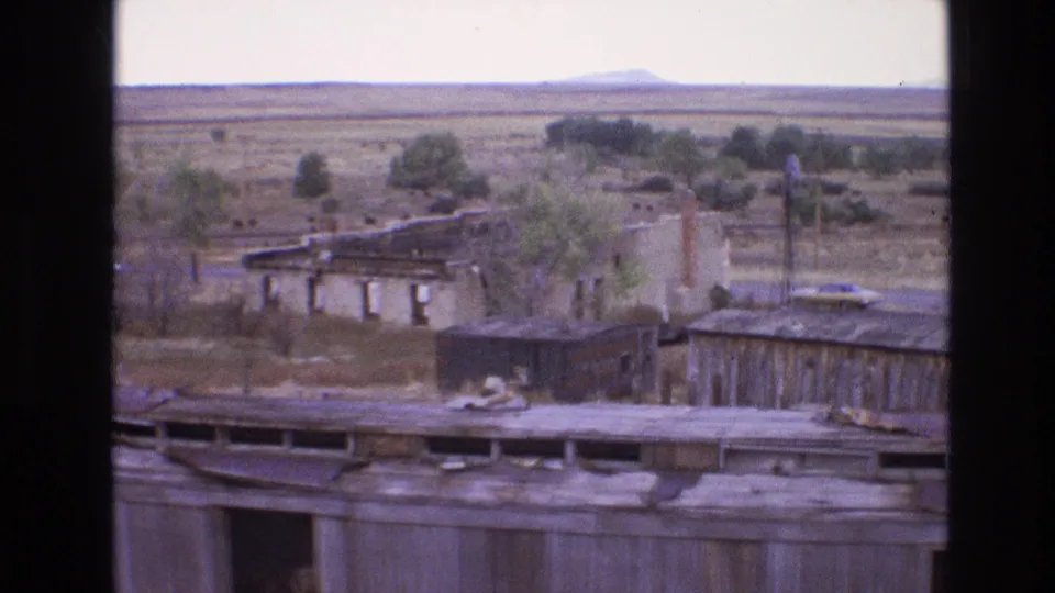 1973 New Mexico home-movie frame of abandoned rail-area buildings with rusted roofing, nearby trees, dry grass, open plains, cattle, and a distant low hill.