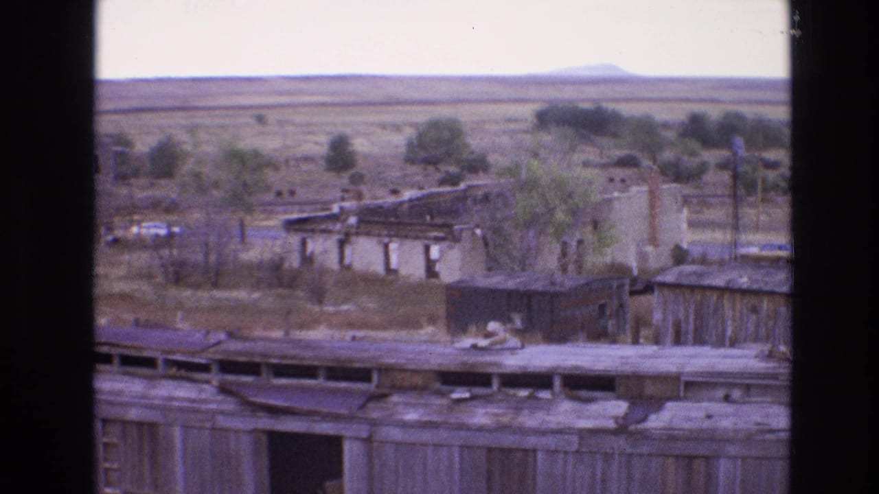 1973 8mm New Mexico frame showing weathered low buildings, scrubby open ground, distant hills, and a cloudy sky.