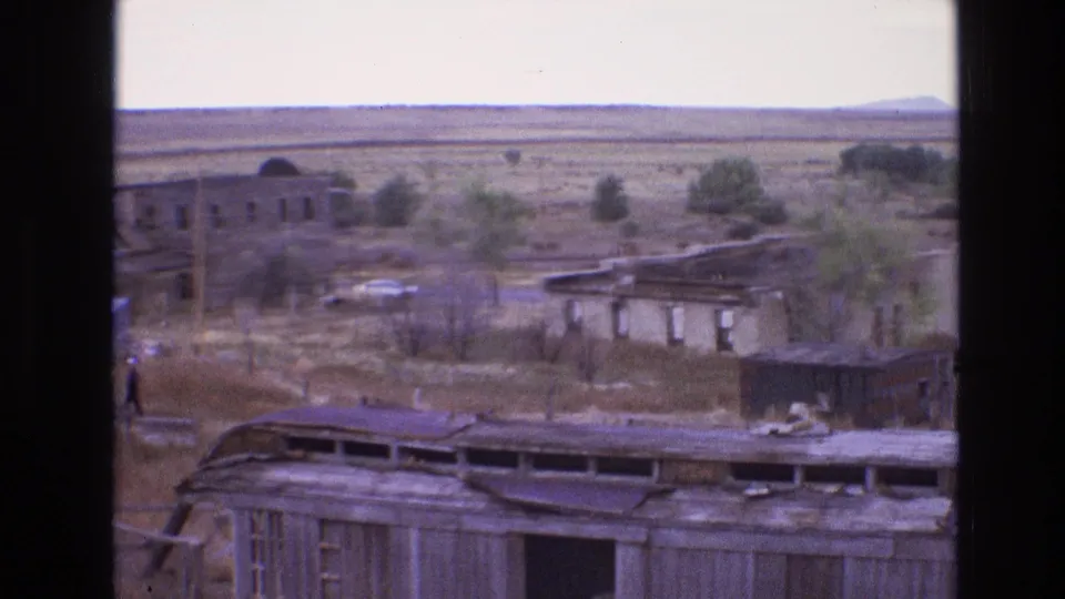 1973 8mm New Mexico frame with ruined wooden and adobe structures, open scrubland, and a distant person walking near the buildings.