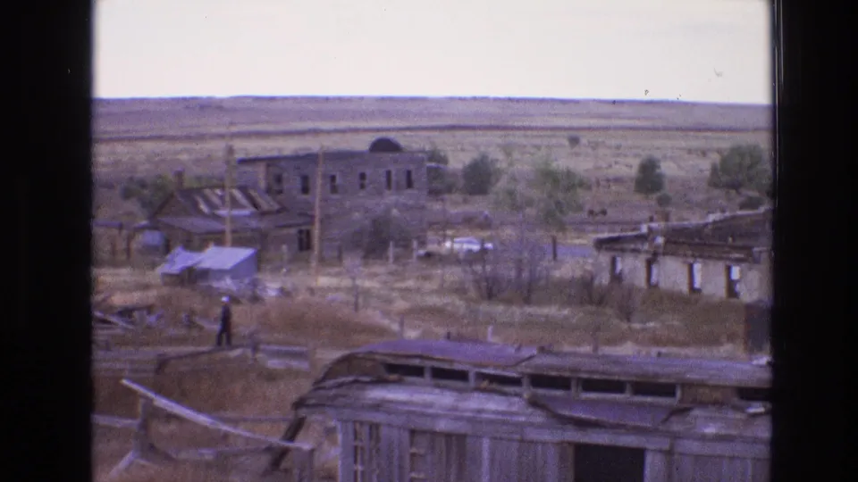 1973 8mm New Mexico frame showing a lone man walking through a ruin-strewn site with damaged buildings, poles, scrub, and open land beyond.