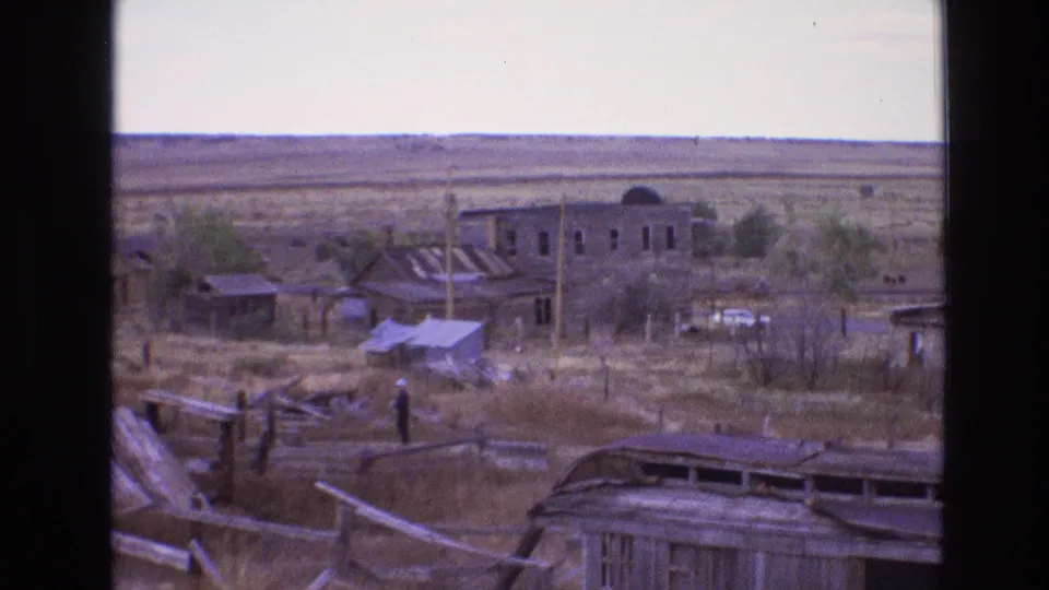 1973 8mm New Mexico frame with a distant man among debris, a damaged roofline in the foreground, ruined buildings, brush, and a broad dry plain.