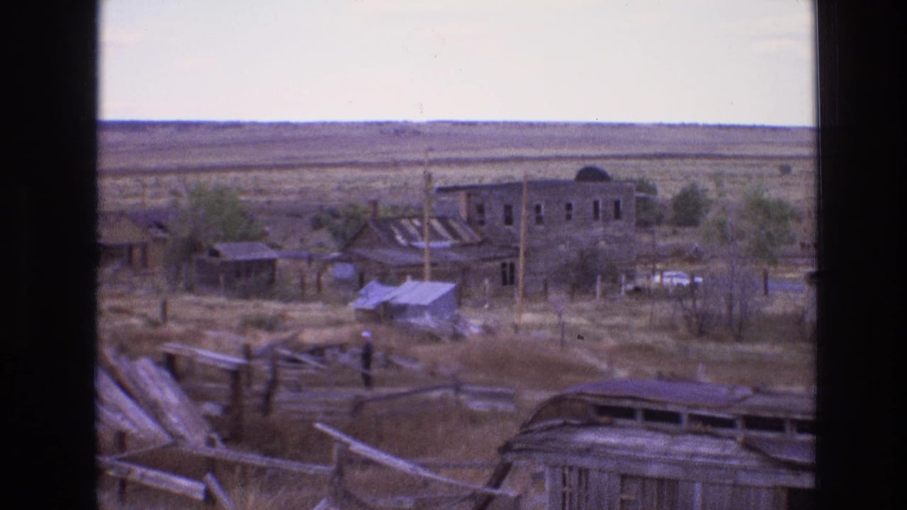 1973 New Mexico 8mm frame showing a weathered desert settlement with rough timber fencing, scattered boards, small structures, utility poles, and open arid terrain.
