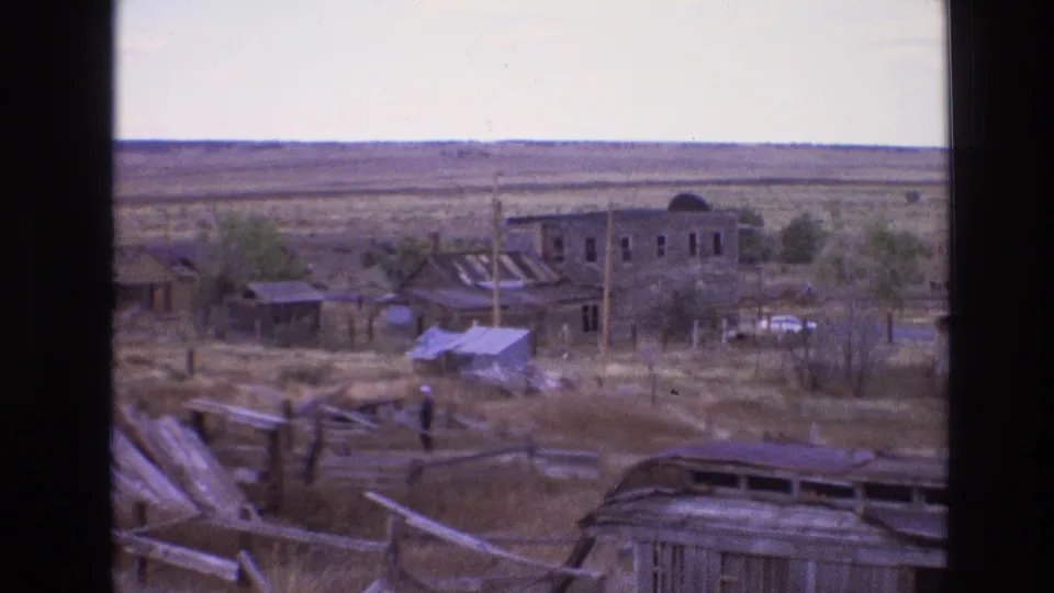 1973 New Mexico 8mm frame showing a weathered desert settlement with rough timber fencing, scattered boards, small structures, utility poles, and open arid terrain.