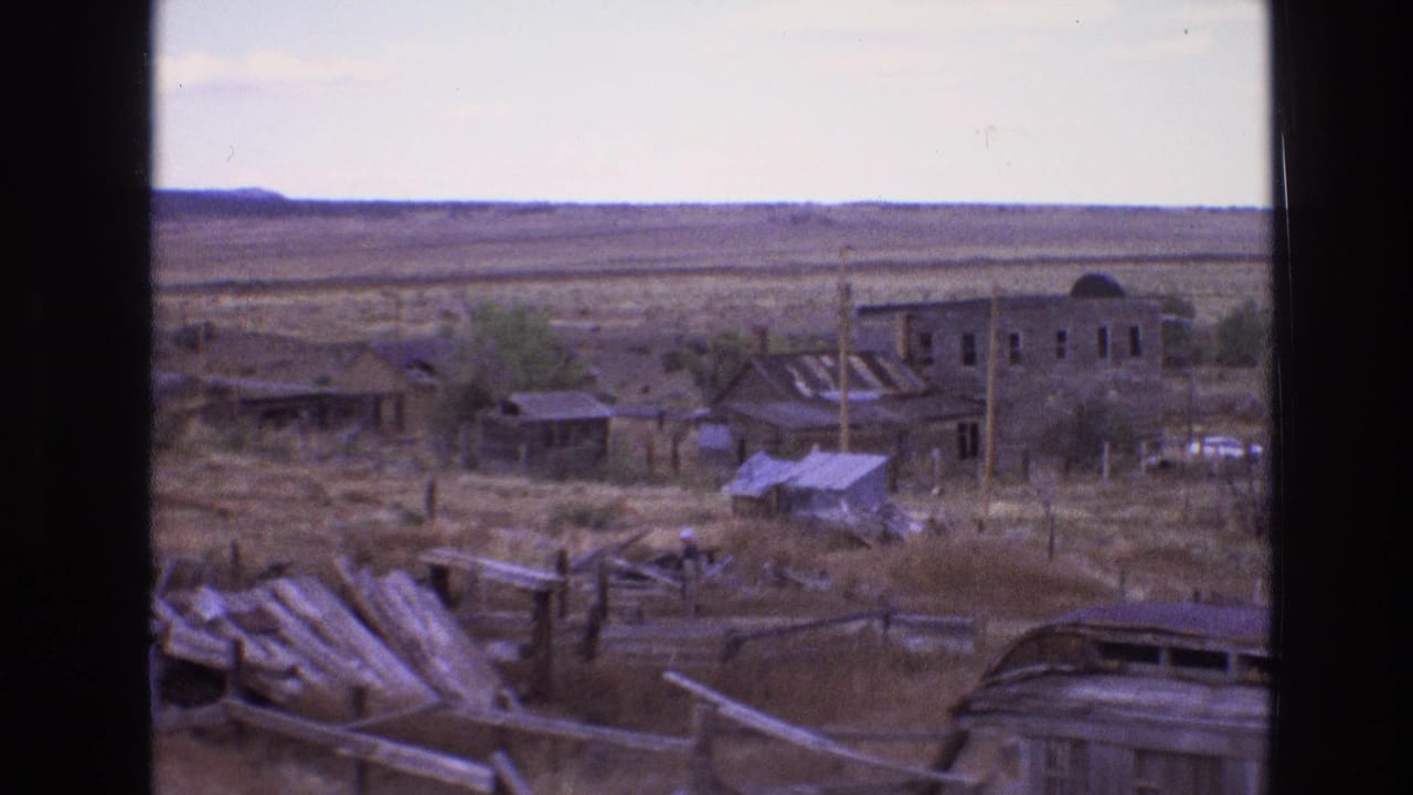 Desert settlement with damaged fencing, low buildings, debris, and a distant unfinished-looking structure.