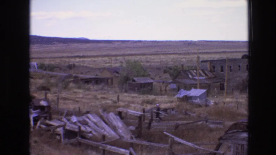 1973 New Mexico 8mm frame showing a man in dark clothing and a light hat walking through a dry settlement with broken fences, debris, and weathered buildings.