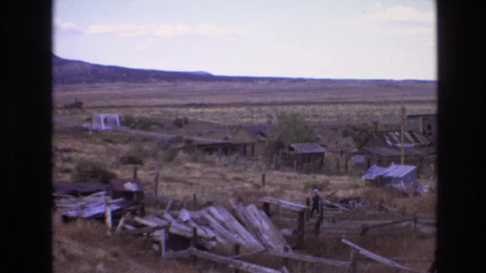 1973 New Mexico 8mm frame of a man walking across an arid settlement with damaged timber fencing, scattered materials, low buildings, utility poles, and distant mesa-like terrain.