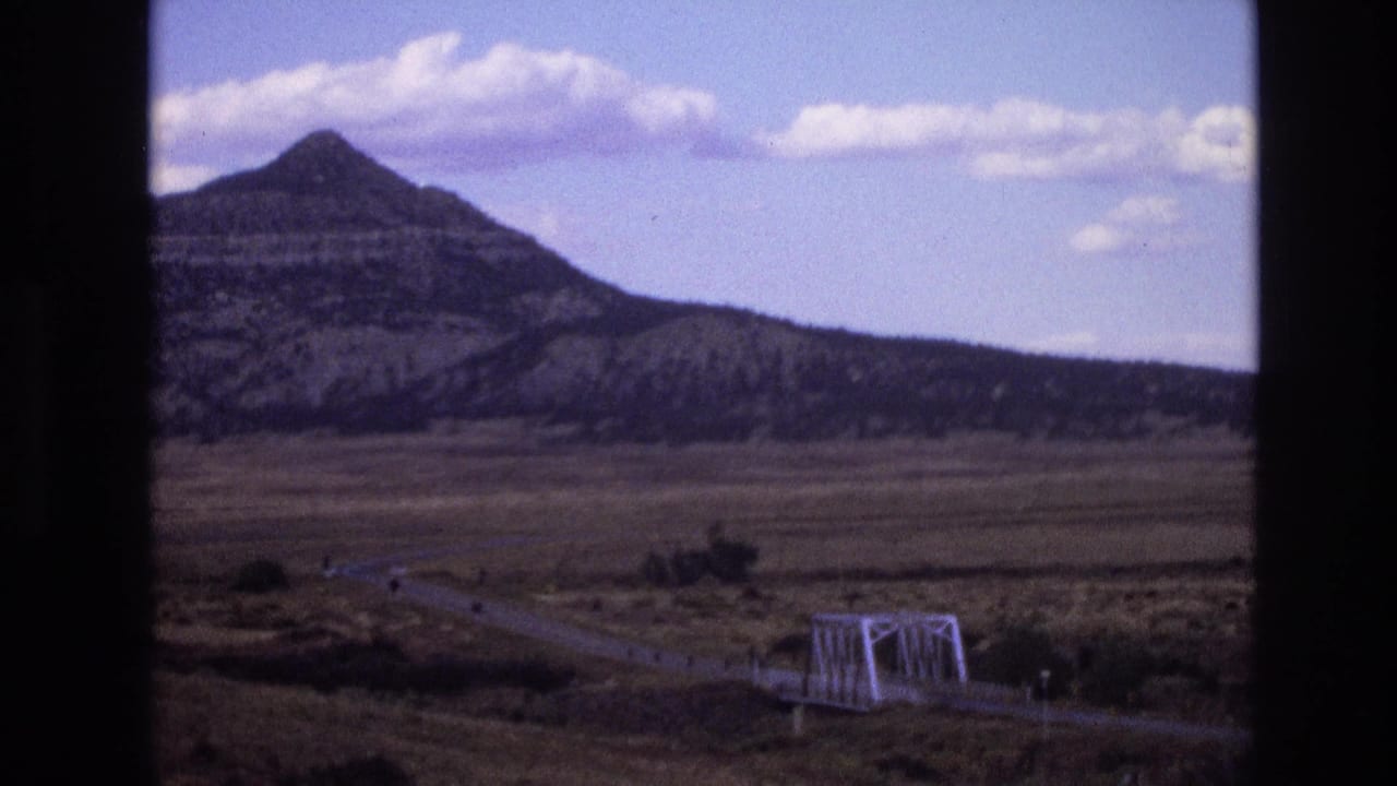 1973 New Mexico 8mm frame showing a layered mountain under scattered clouds, with an open arid plain, curving road, and white truss bridge in the lower foreground.