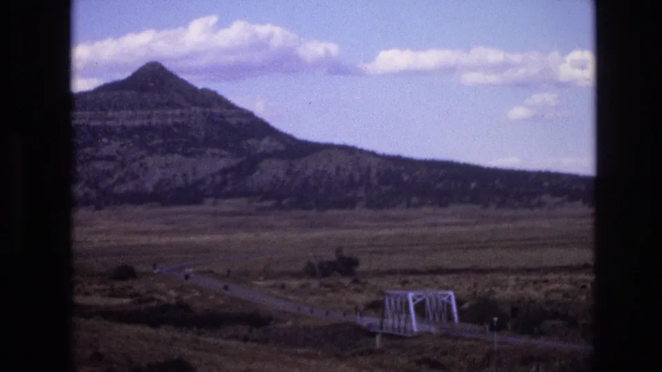 1973 New Mexico 8mm frame showing a layered mountain under scattered clouds, with an open arid plain, curving road, and white truss bridge in the lower foreground.