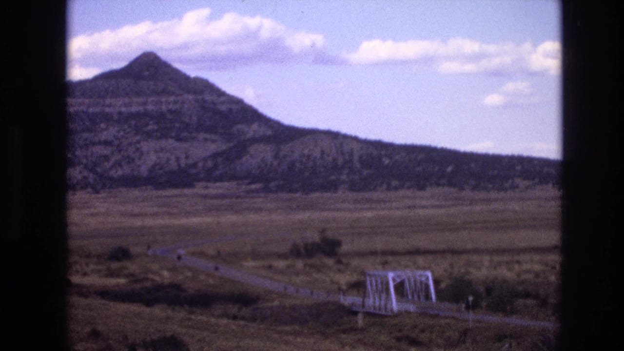 Mountain and clouds over a dry plain with a white bridge and road near the lower edge of the frame.
