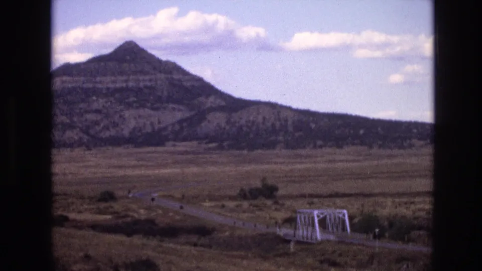 1973 New Mexico 8mm frame showing a distant layered mountain, dry open land, a rural road, and a small white truss bridge crossing a low channel.