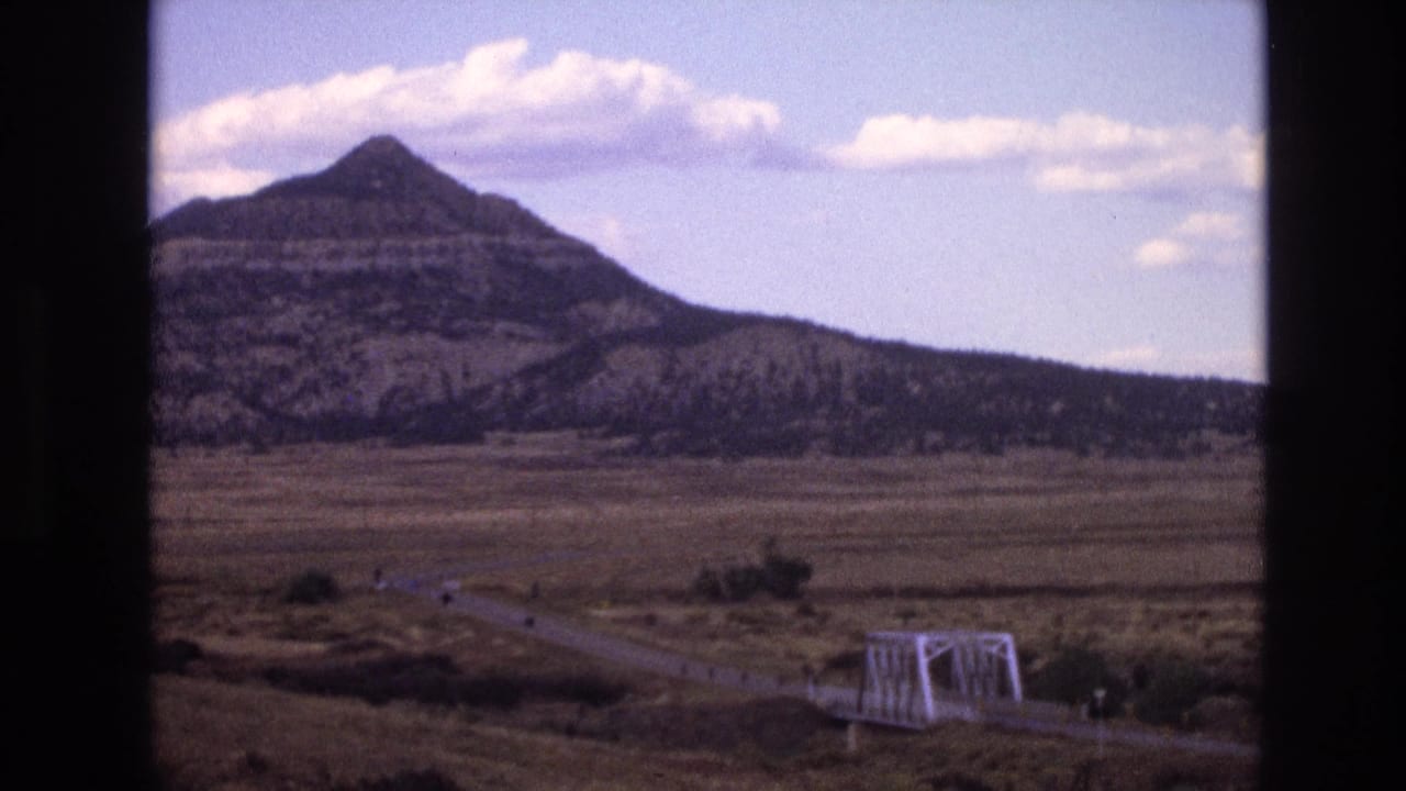 1973 8mm home-movie frame from New Mexico showing a winding rural road, small white truss bridge, broad plain, and layered mountain under a blue sky.