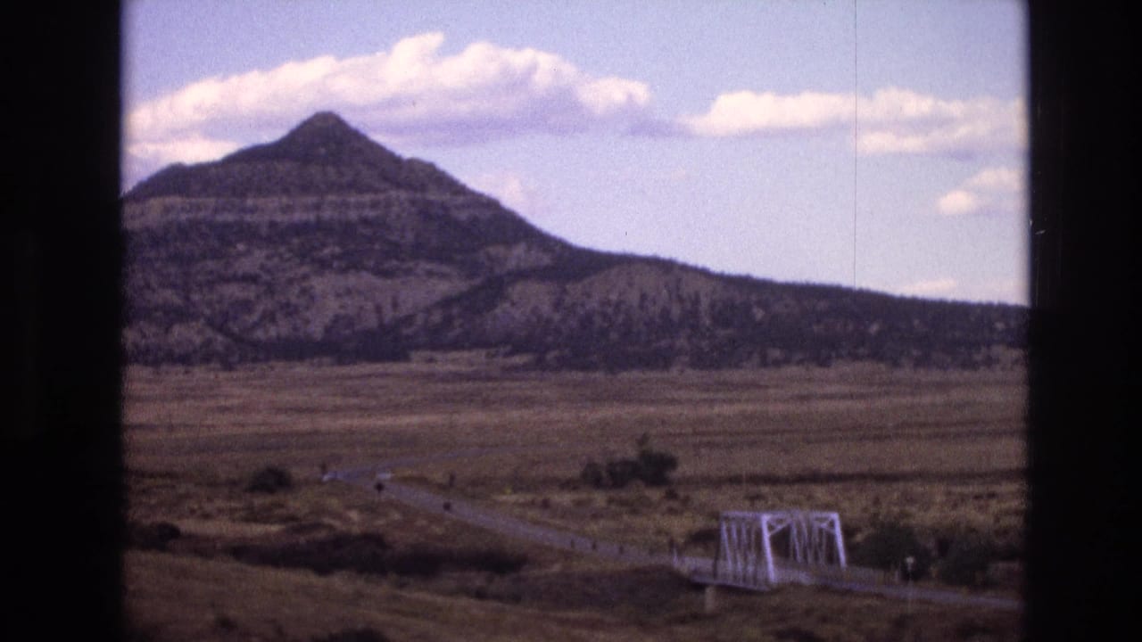 Wide rural plain with a road, small bridge, mountain, and clouds framed by dark film edges.