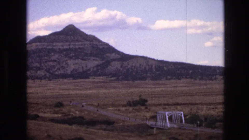 1973 New Mexico home-movie frame with a distant country road crossing a small white bridge before a prominent layered mountain and pale clouds.