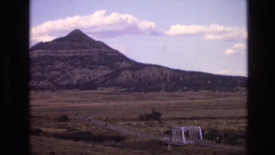 Faded 8mm frame from 1973 New Mexico showing an open plain, winding road, small bridge, and distant layered mountain beneath scattered clouds.