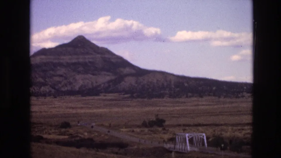 1973 8mm New Mexico frame showing a sparse rural plain, small white truss bridge, curving road, and dark layered mountain with cloud cover above.