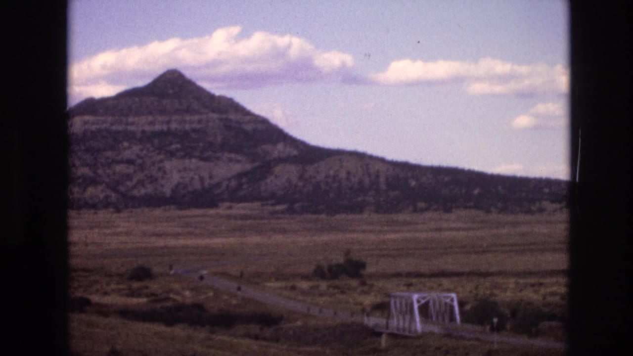 1973 New Mexico 8mm frame showing a mesa beyond dry fields, a rural road, and a small white truss bridge under a cloudy sky.