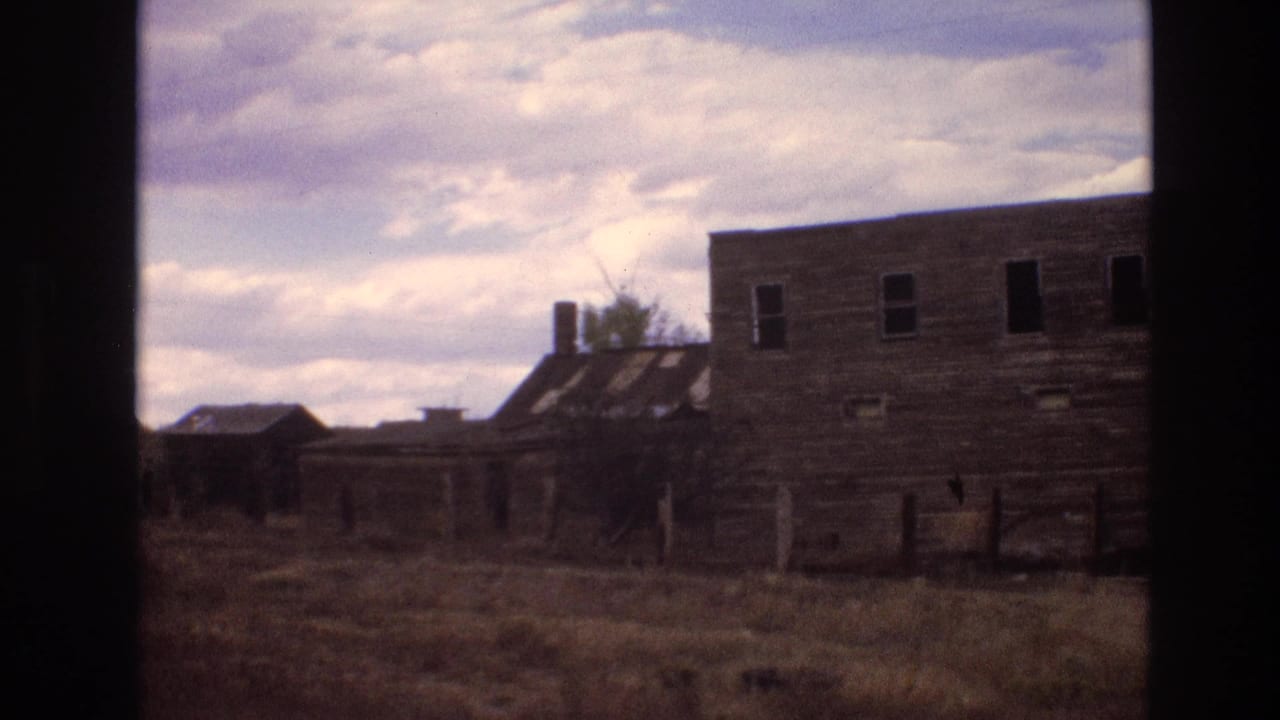 1973 New Mexico 8mm frame showing a weathered wooden building with dark window openings beside dry grass and distant mesa terrain.