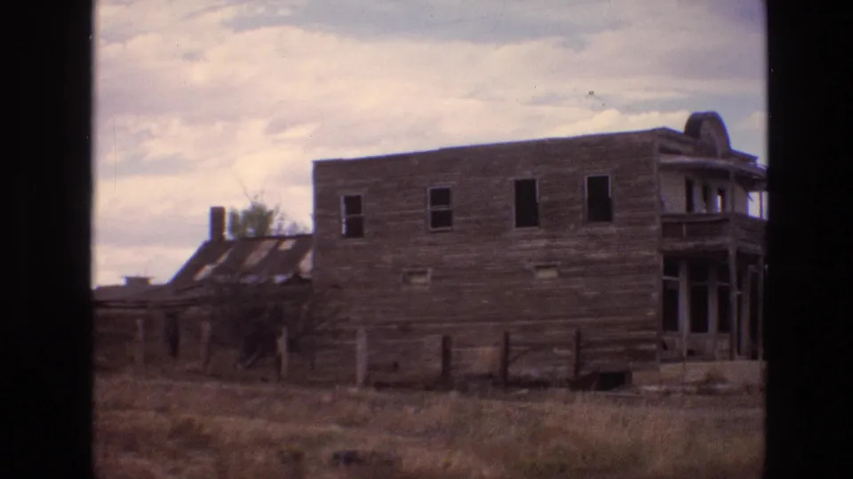 1973 New Mexico 8mm frame showing the side of a weathered wooden structure with dark window openings, fence posts, and dry field grass.
