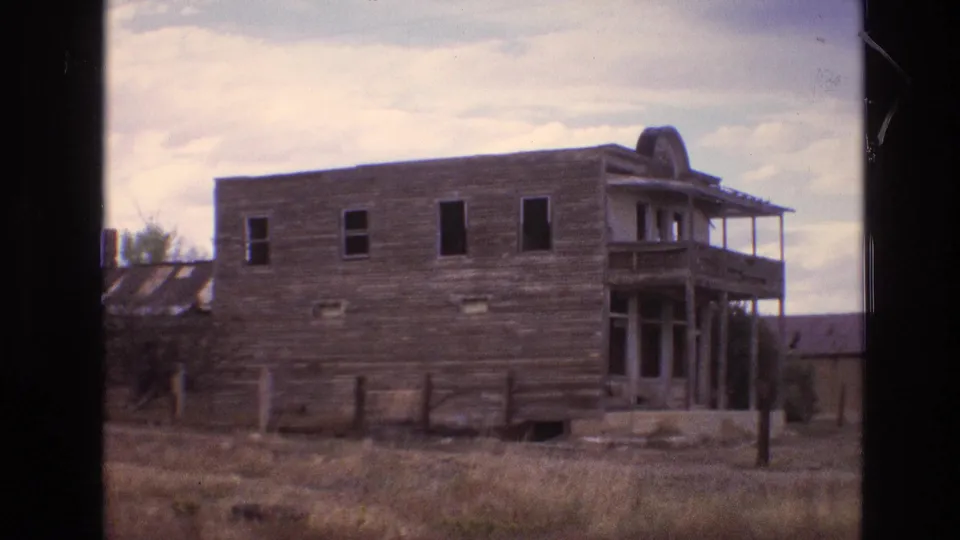 1973 New Mexico 8mm frame showing a weathered wooden building with porch supports, open dark windows, fence posts, and dry grass under a pale cloudy sky.