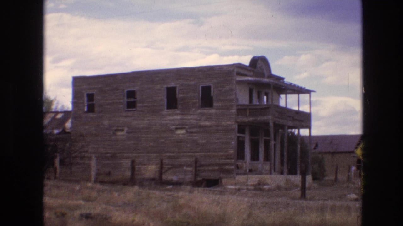 1973 New Mexico 8mm frame showing a weathered two-story wooden building with dark window openings, porch structure, fence posts, and dry roadside grass.