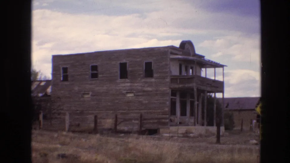 1973 New Mexico 8mm frame showing a weathered two-story wooden building with dark window openings, porch structure, fence posts, and dry roadside grass.