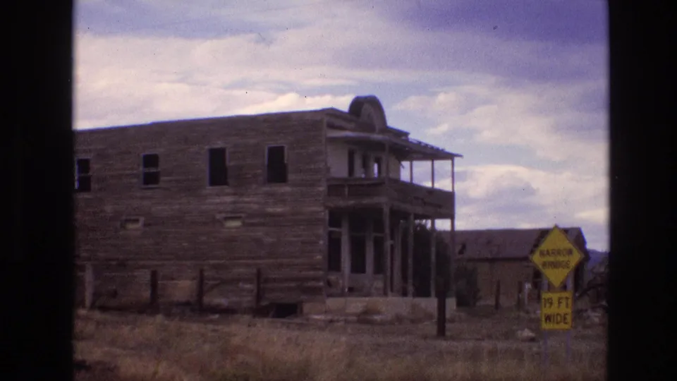 1973 New Mexico 8mm frame with the abandoned-looking wooden building, porch, dry grass, outbuilding, and a yellow sign reading narrow bridge 19 ft wide near the roadside.