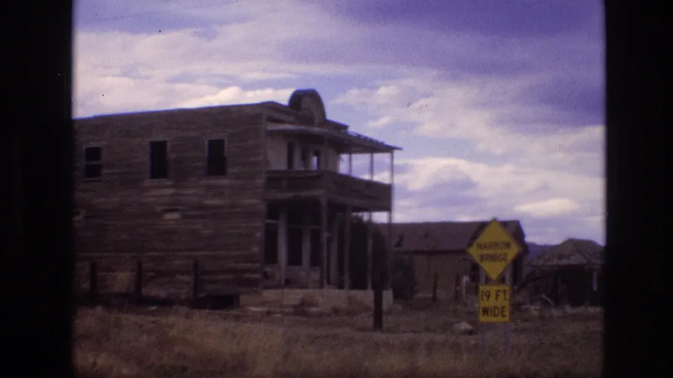 1973 New Mexico 8mm frame showing the panned view with the weathered building partly cropped, a yellow narrow bridge sign in front, dry grass, and low structures beyond.