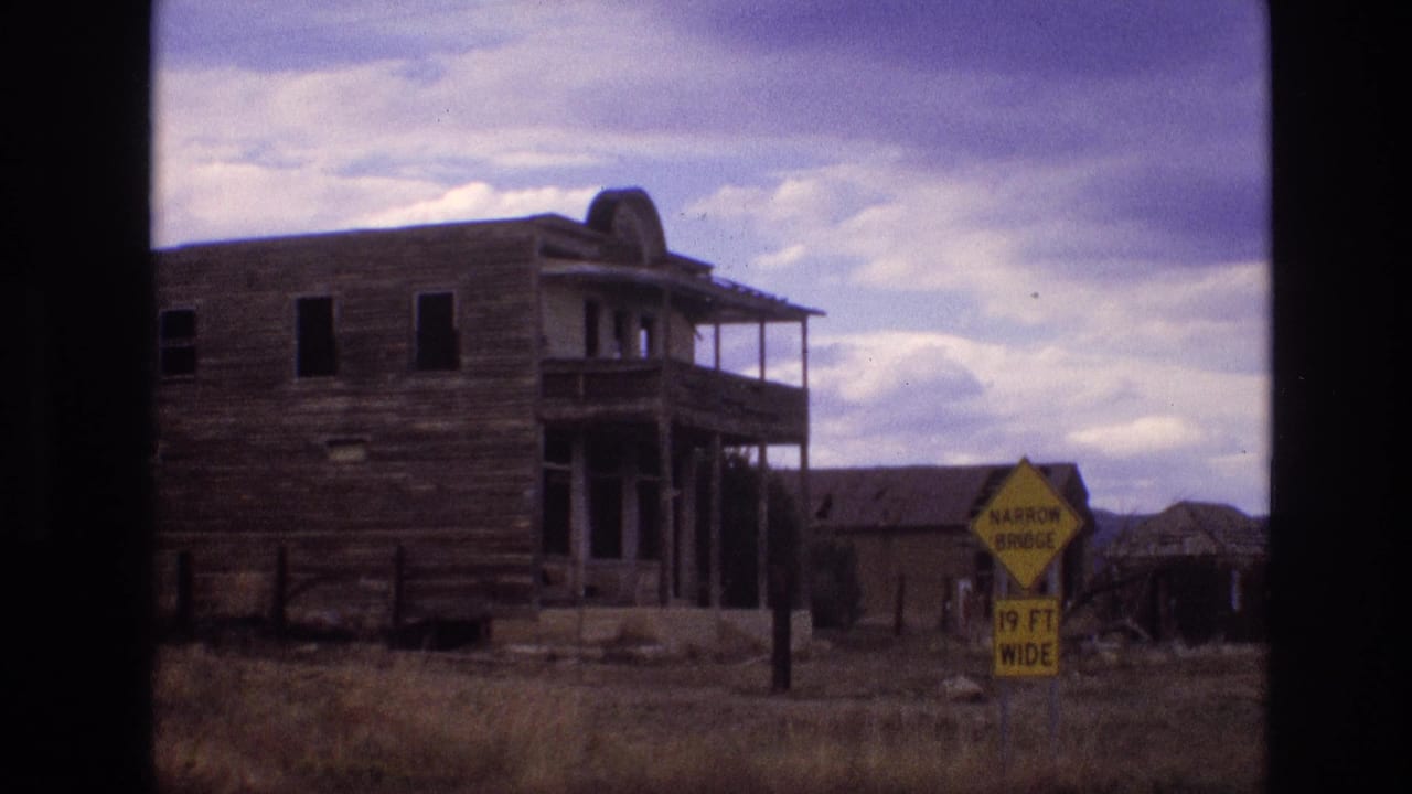 1973 New Mexico 8mm frame with a weathered two-story wooden building, porch structure, and yellow narrow bridge sign beside a rural road area.