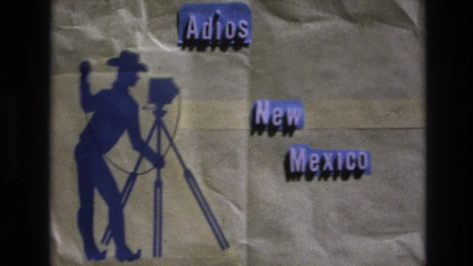 Soft 8mm frame from the 1973 New Mexico reel showing a handmade "Adios New Mexico" card, blue cowboy figure, tripod camera, textured paper, and visible projection or scan edges.